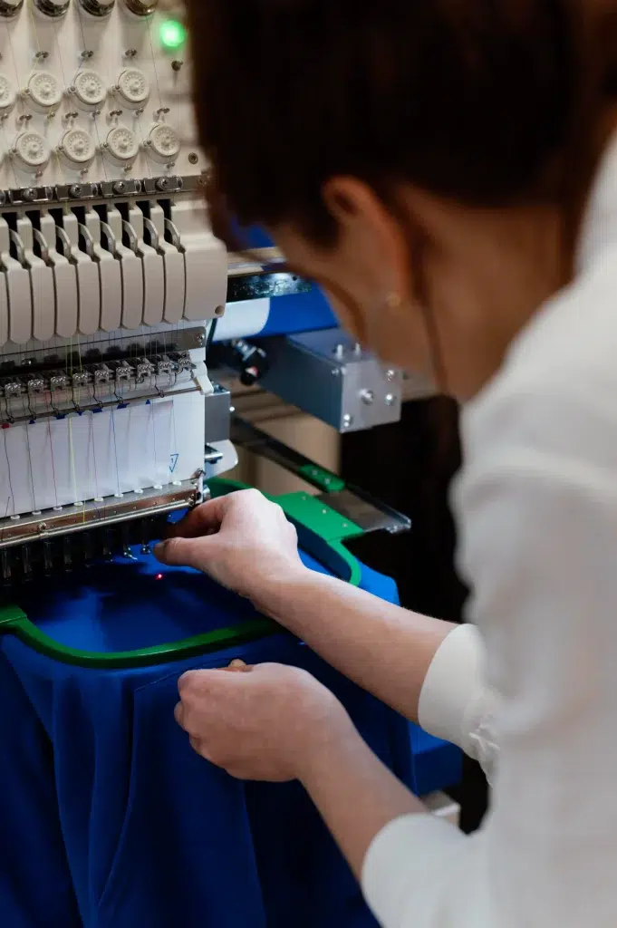 Une personne ajuste un tissu bleu sur le cadre d'une machine à broder, avec de nombreuses aiguilles et bobines de fil visibles.